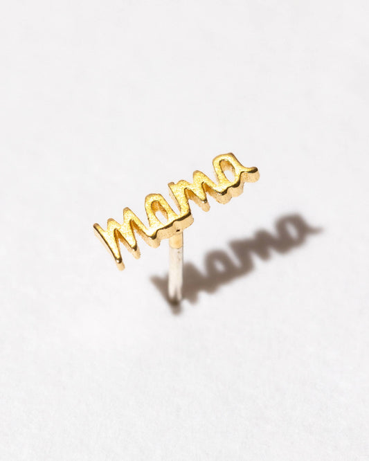 A single gold-colored earring with the word 'MAMA' written in a stylized font, placed against a white background.