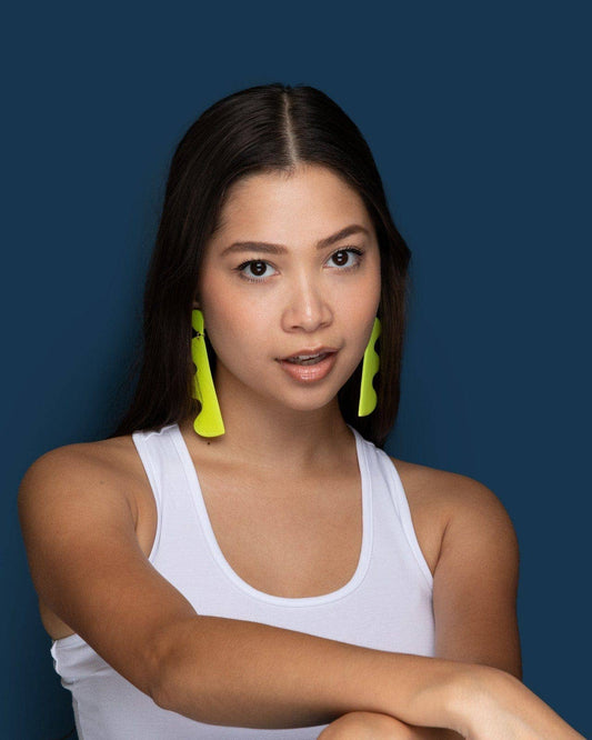 Woman wearing a white tank top and bright green earrings against a dark blue background