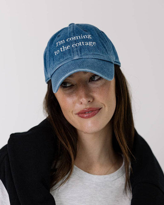 Woman wearing a blue cap that says "I'm coming to the cottage" and a black jacket over a white shirt with graphics.