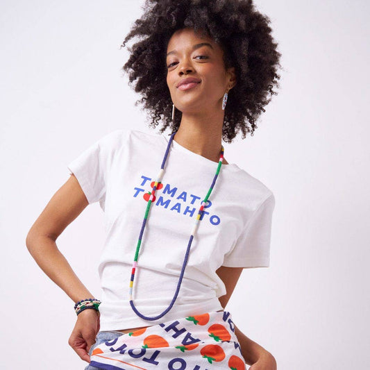 Person wearing a white t-shirt with colorful text and a skirt with fruit pattern and a colorful beaded necklace on a plain background