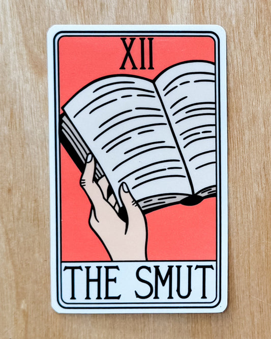 A sticker with a tarot card design featuring the number 12, a hand holding an open book, and the word 'THE SMUT' in bold letters.