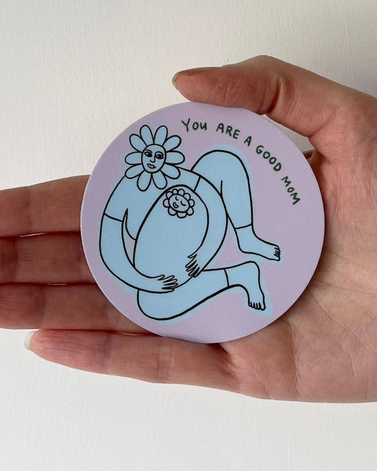 Hand holding a circular sticker that has an illustration of a mom and baby and says: You are a good mom.