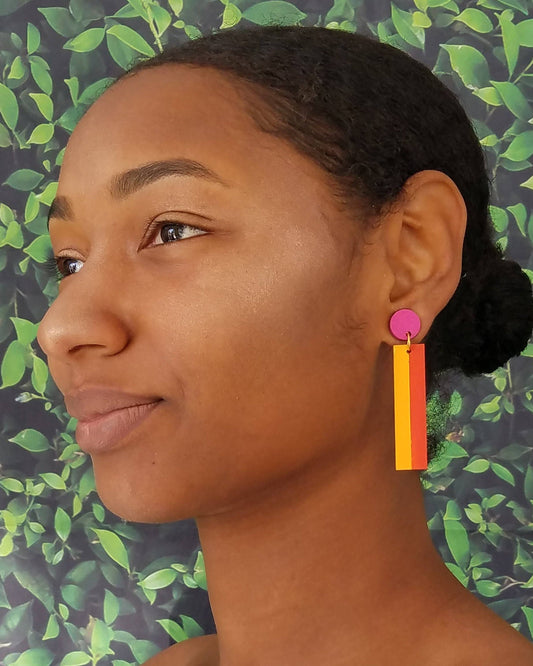 A woman shown in profile wearing a bright pink circular earring with a long rectangular drop charm in pink and orange, set against a green leaf patterned background.
