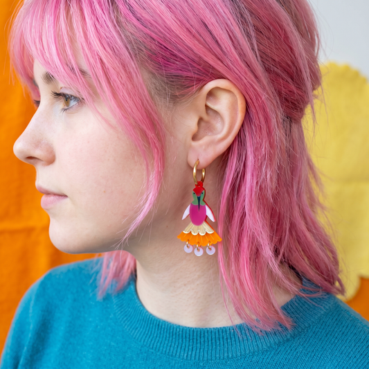 A close-up side profile of a person with bright pink hair wearing a colorful dangling earring. The earring has a gold hoop and a tiered charm in pink, white, orange, green, and red with small dangling beads at the bottom.