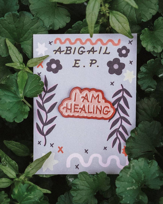 Small enamel pin that says 'I AM HEALING' on a bed of green leaves.
