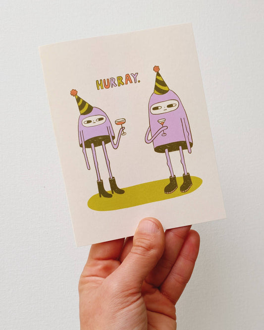 Hand holding a card with cartoon characters and 'HURRAY!' text against a plain background