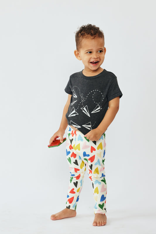 A toddler wearing Lilla Barn Clothing organic leggings with a multicolored heart pattern, paired with a black top featuring a heart graphic.