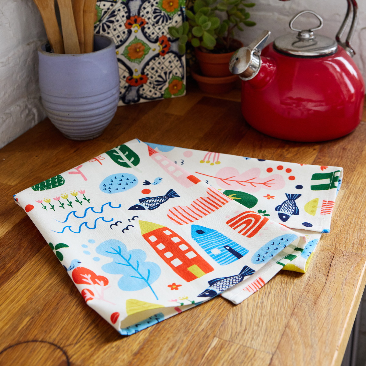 white organic cotton tea towel with brightly colored illustrations on it sitting on a wooden counter by a tea pot