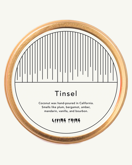 A circular travel tin candle with a label that reads 'Tinsel' and lists the scents such as coconut, vanilla, and bourbon. It also mentions 'California, USA' and 'hand-poured in California, USA'.