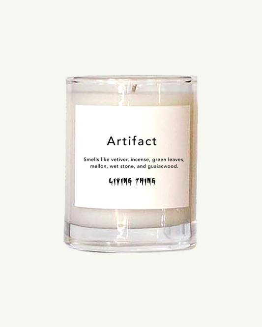 A clear glass votive candle with a white label that reads 'Artifact' and lists the scents including vetiver, melon, wet stone, and guaiacwood. It also mentions the coconut wax and cotton wick.