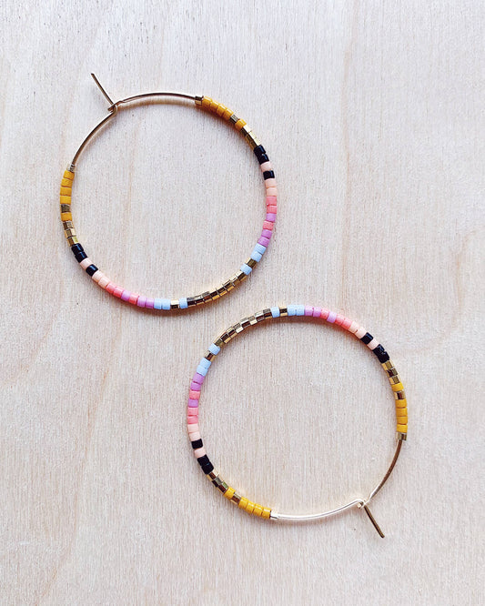 A pair of seed bead hoop earrings with gold-fill hoops and multicolored beads, displayed on a wooden surface.