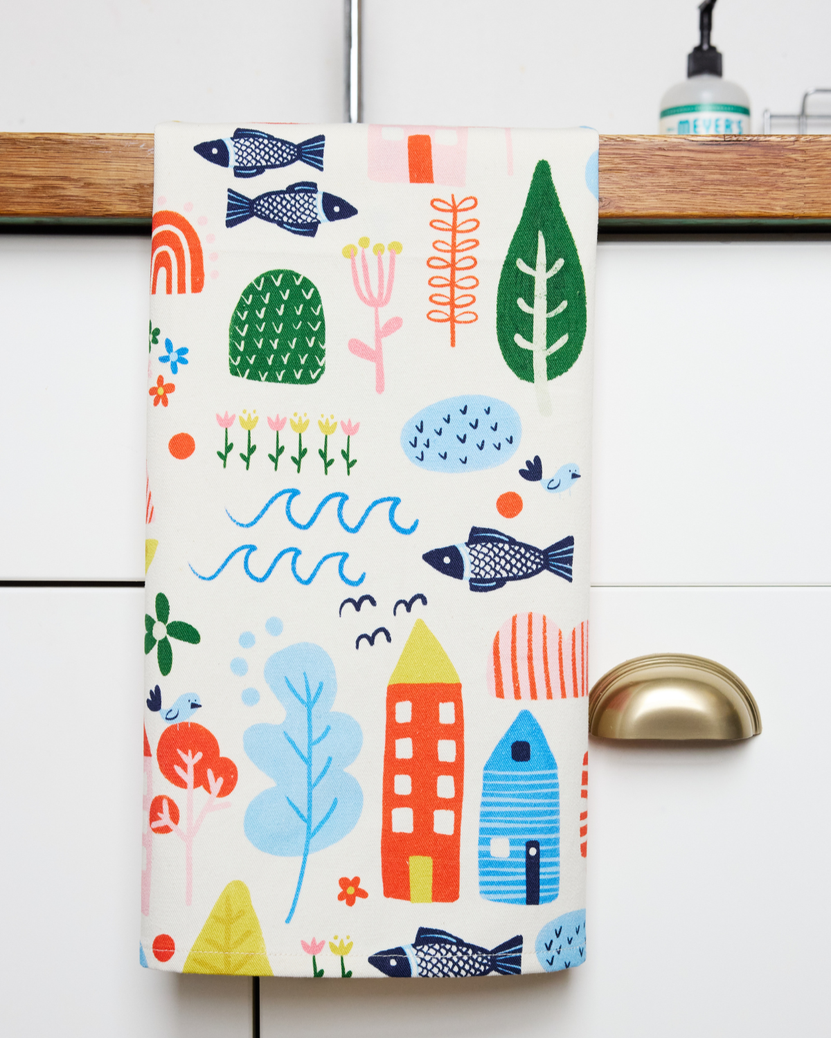 cream kitchen towel with colorful folk art illustrations including a house, leaf, waves, fish, flowers, and more