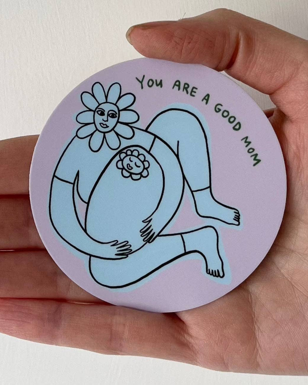 Hand holding a circular sticker that has an illustration of a mom and baby and says: You are a good mom. 
