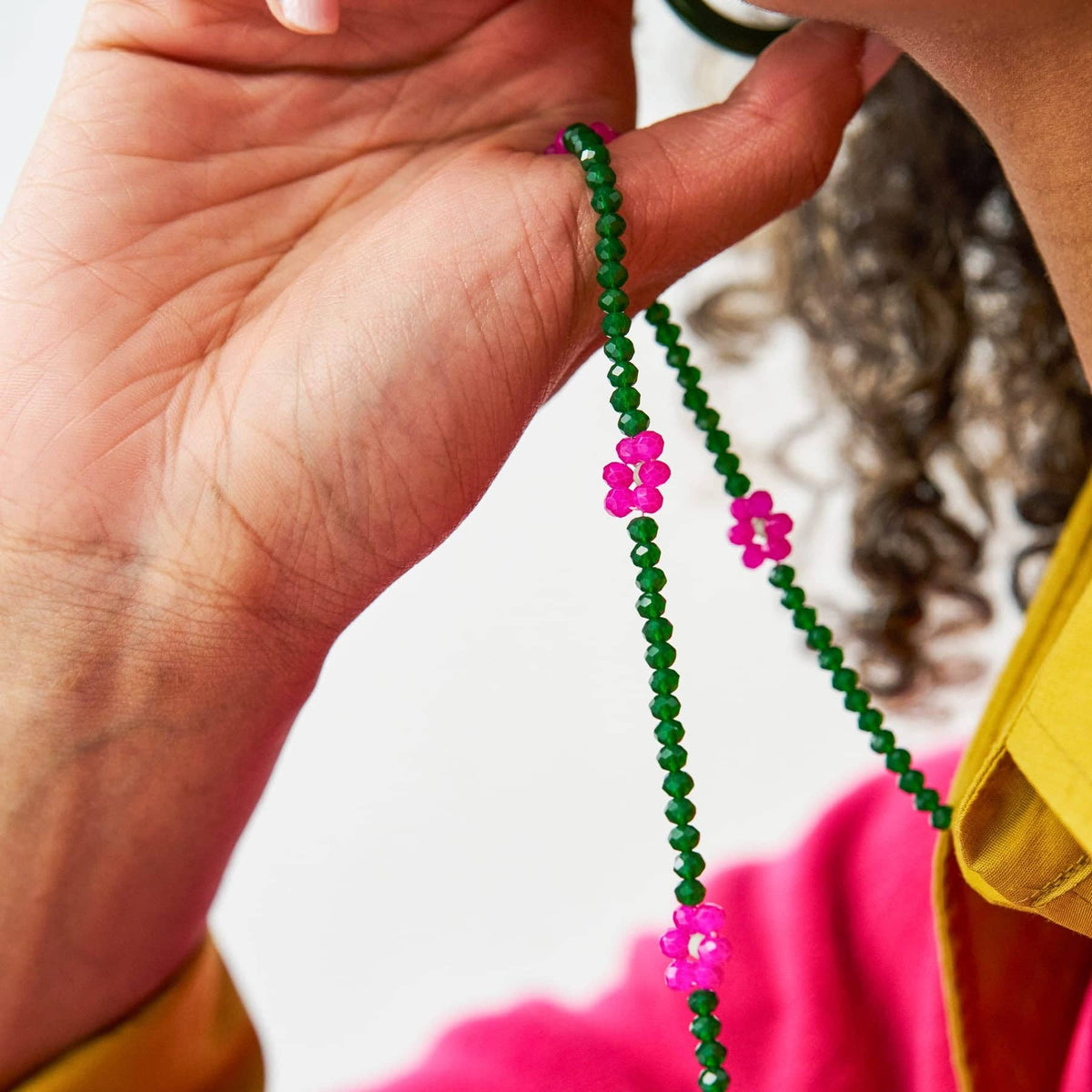 A close-up of a person holding a green beaded necklace with pink flower-shaped beads against a bright white background.