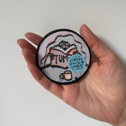 Hand holding a round embroidered patch with a person reading a book design on a white background
