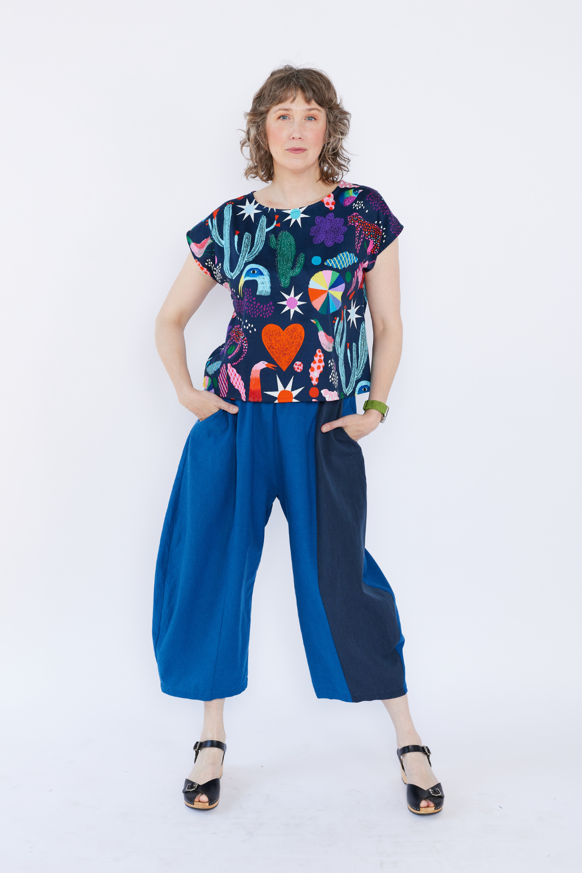 Woman wearing a colorful Lilla Barn top with plant pattern and blue pants on a white background