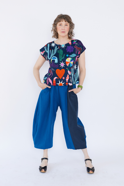 Woman wearing a colorful Lilla Barn top with plant pattern and blue pants on a white background