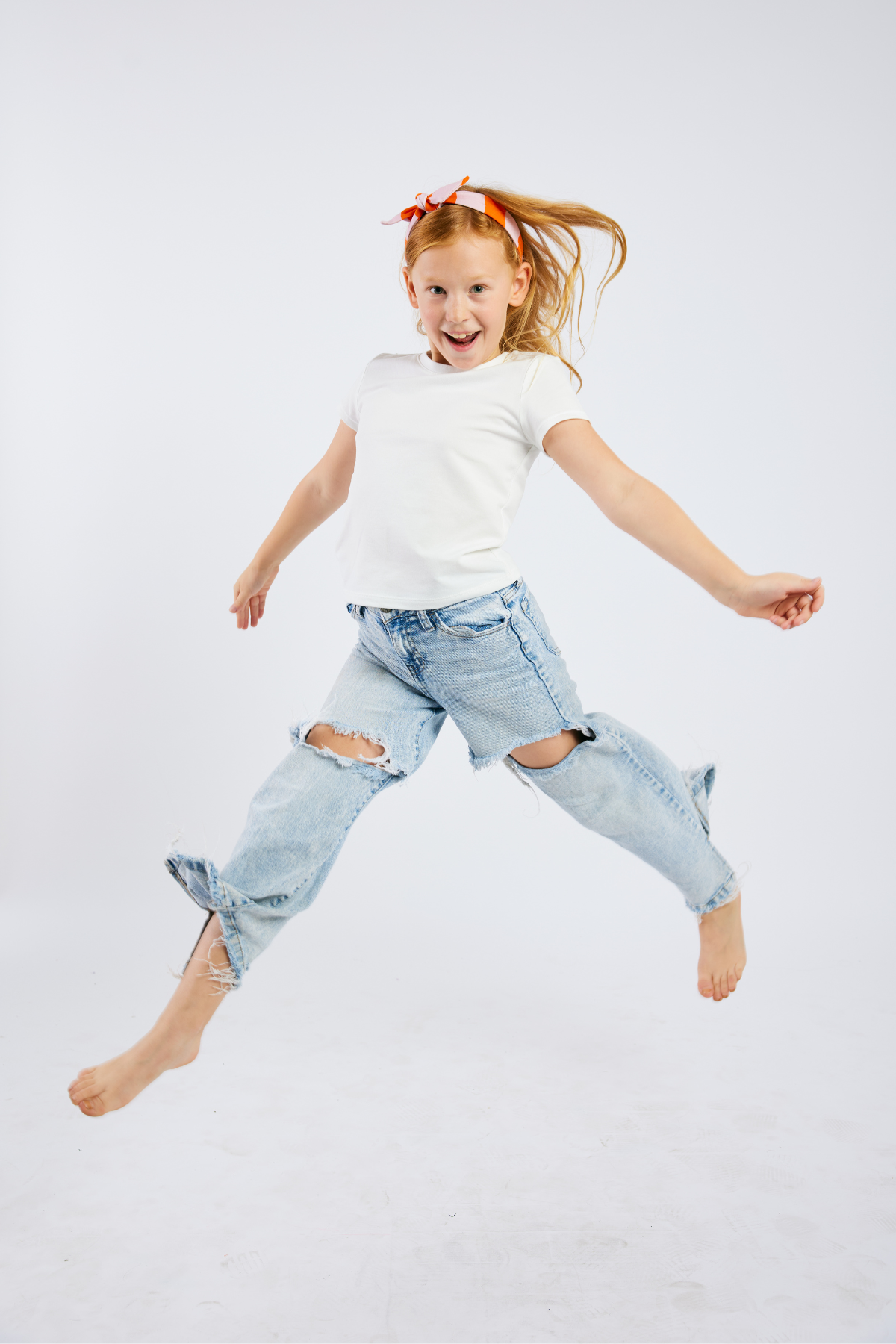 child jumping in the air wearing  jeans, a white top, and a pink and orange striped stretchy headband