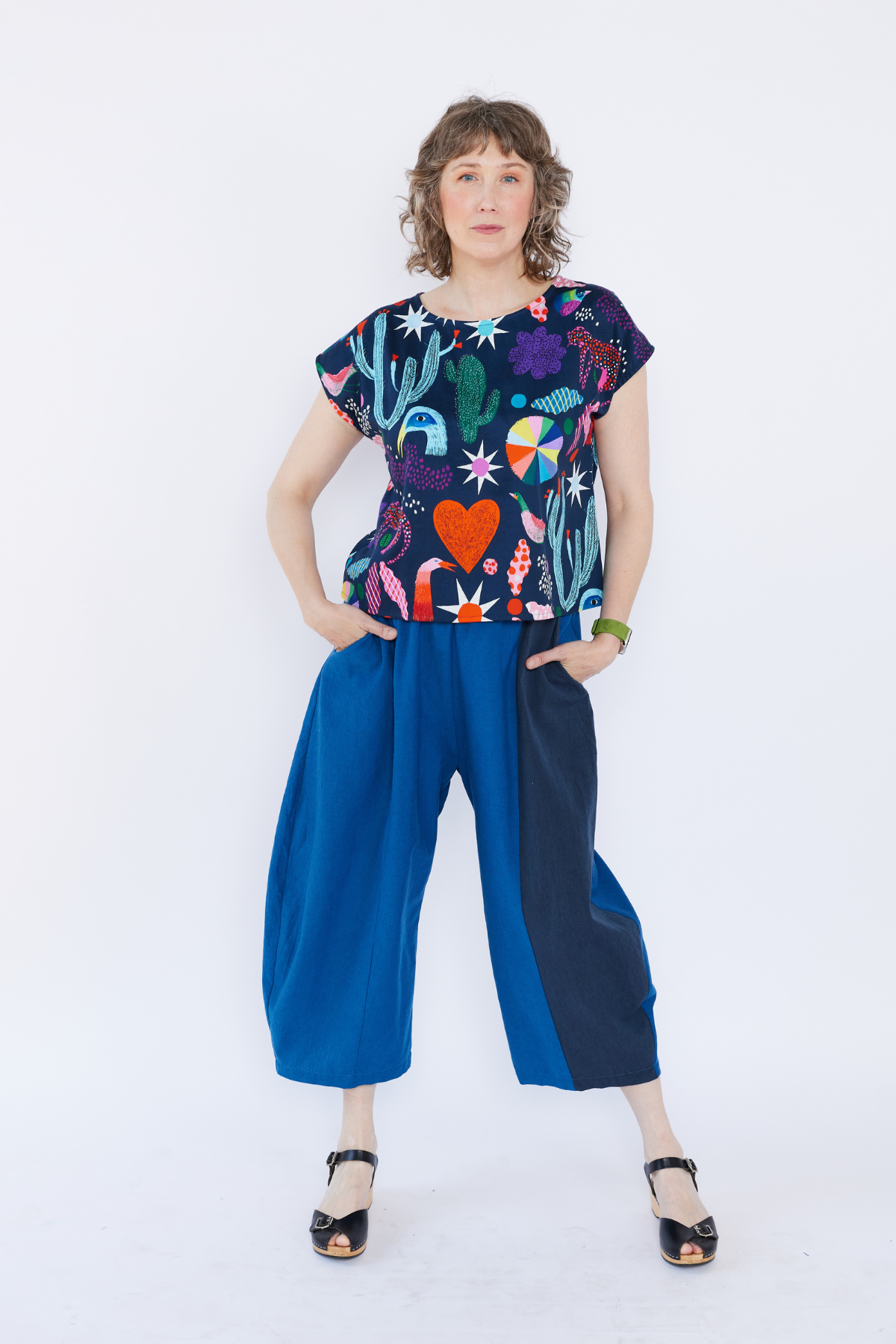 Woman wearing a colorful top with cactus and heart designs and blue pants on a white background