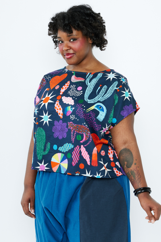 Person wearing a colorful patterned top by Lilla Barn Clothing with blue pants on a white background
