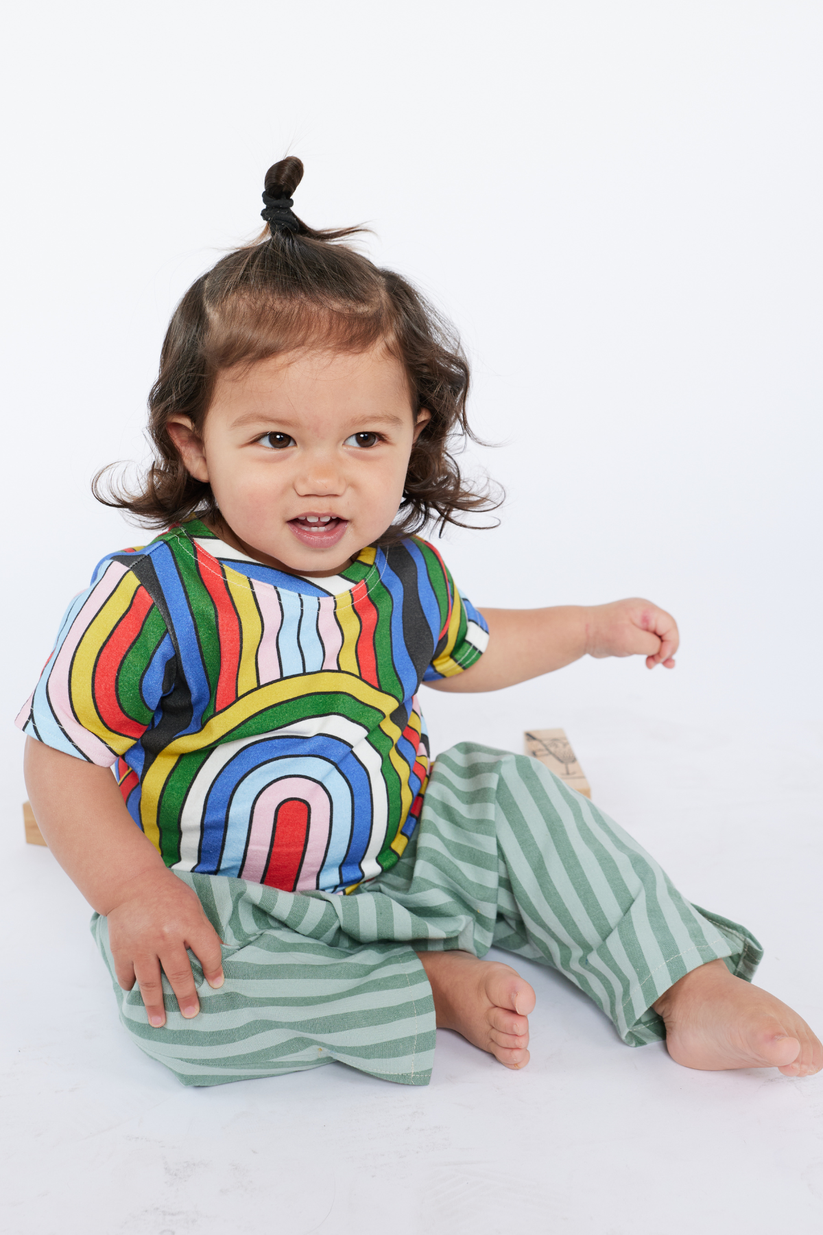 Child wearing a colorful rainbow shirt and green striped pants from Lilla Barn Clothing on a white background