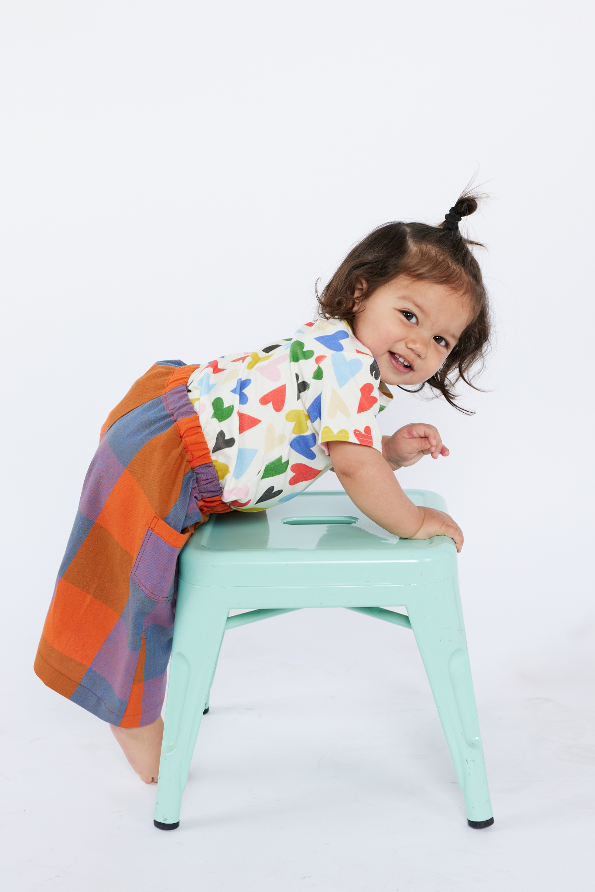 Child wearing a colorful shirt and rainbow-patterned pants on a light blue stool against a white background