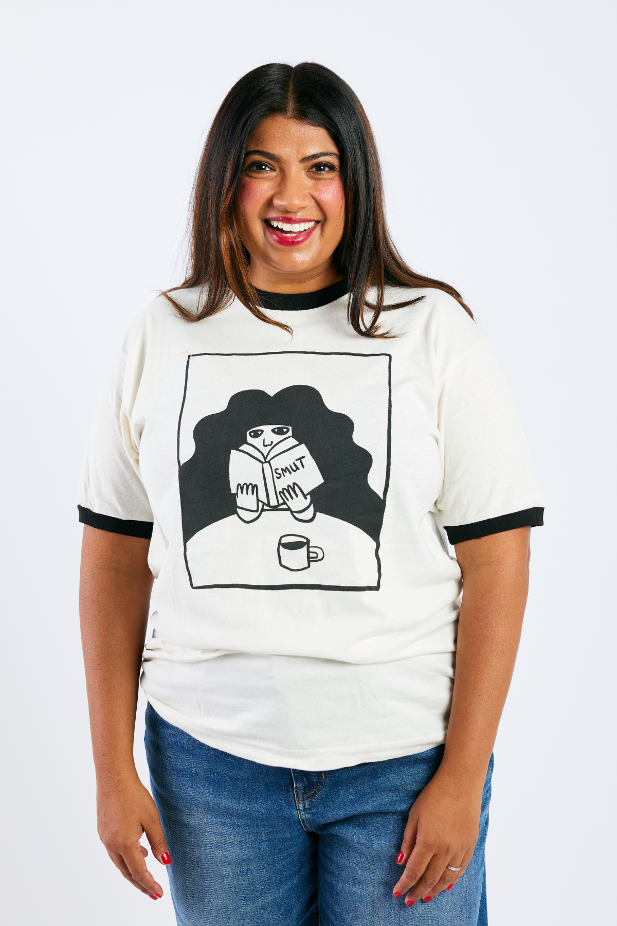 Woman wearing a white t-shirt with an illustrated woman reading smut and drinking coffee on a plain background