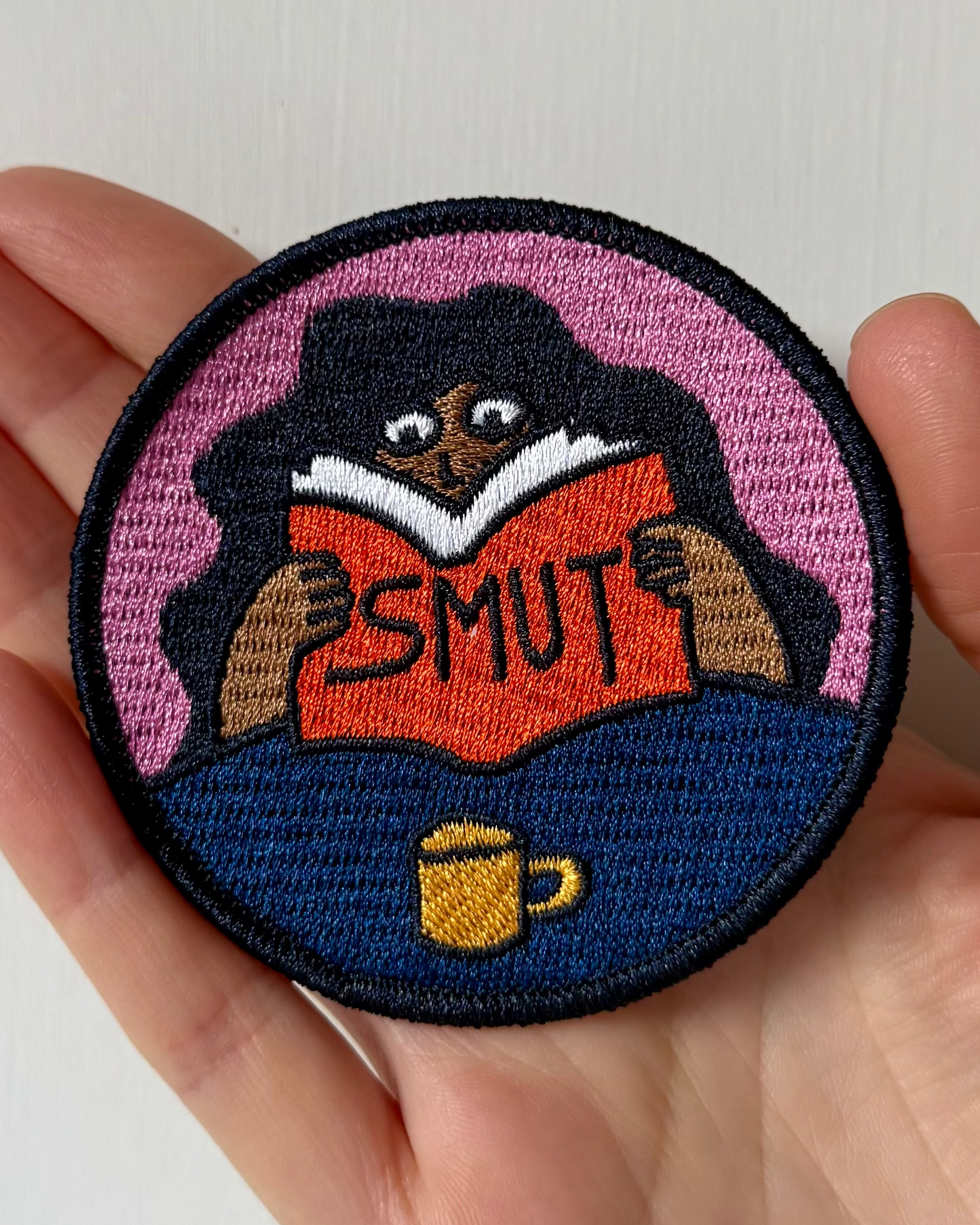 Embroidered patch of a cartoon character reading 'SMUT' with a cup, held in a hand against a white background.