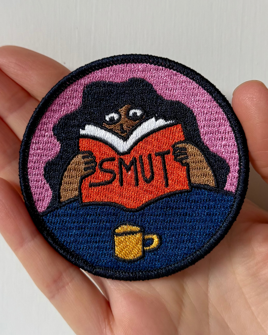 Embroidered patch of a cartoon character reading 'SMUT' with a cup, held in a hand against a white background.