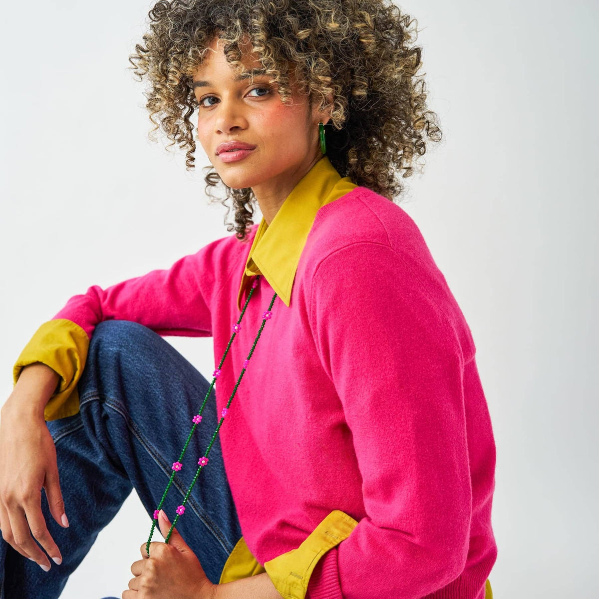 A woman with curly brown hair wearing a bright pink knit sweater with contrasting yellow collar and cuffs, paired with blue jeans, posing against a light gray studio background.