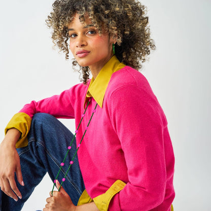 A woman with curly brown hair wearing a bright pink knit sweater with contrasting yellow collar and cuffs, paired with blue jeans, posing against a light gray studio background.