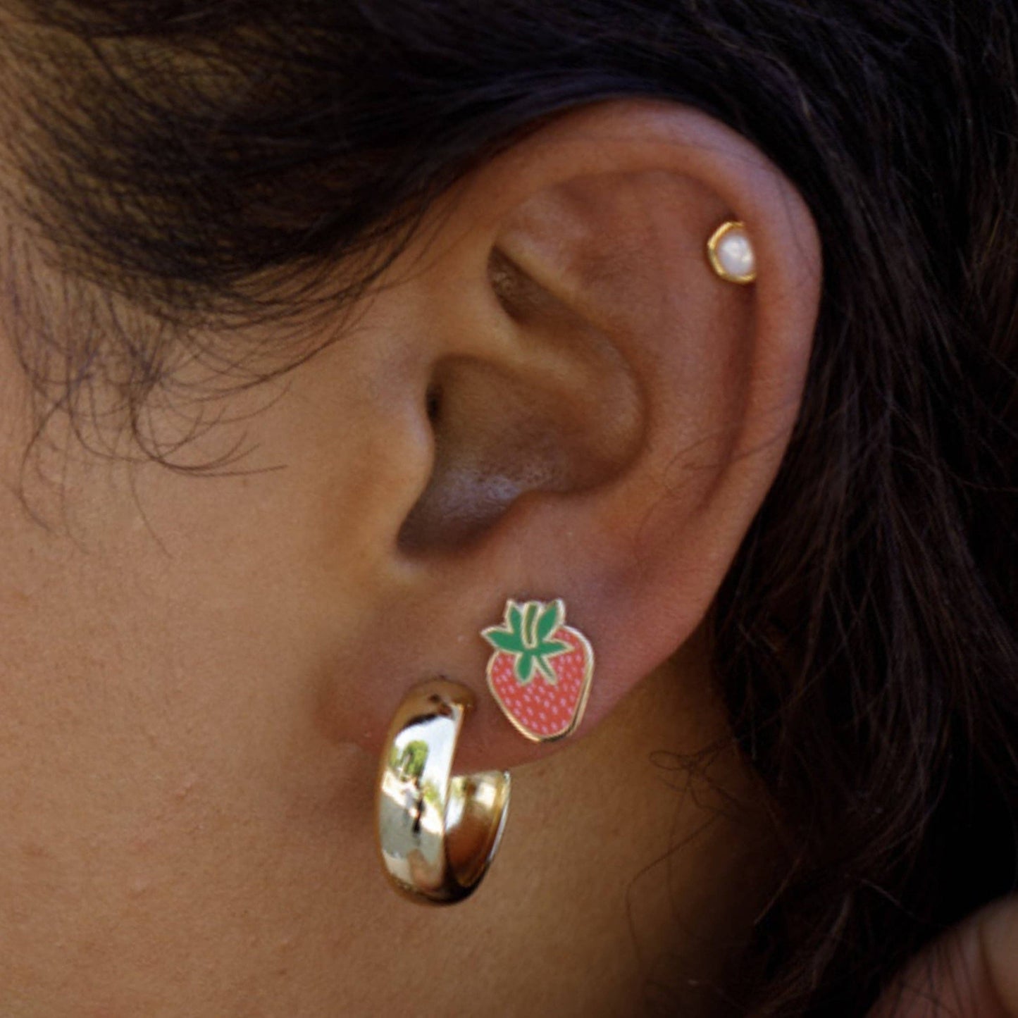 Close-up of an ear with gold hoop earrings and a strawberry-shaped earring.