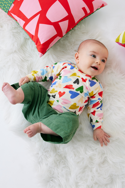 Baby wearing a colorful heart-patterned shirt and green cotton striped pants lying on a white surface with a red pillow in the background.