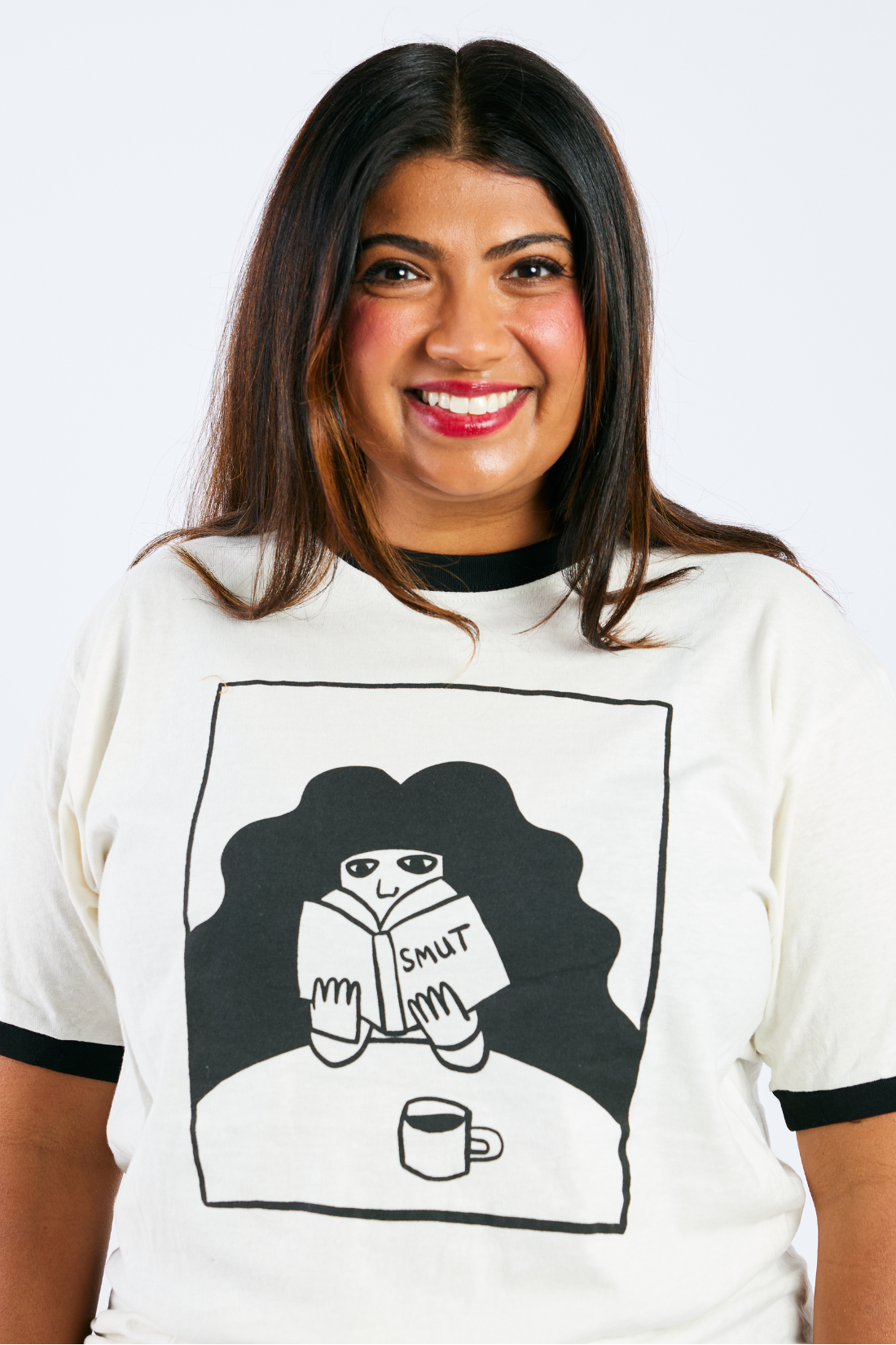 Woman wearing a white t-shirt with an illustrated woman reading smut and drinking coffee on a plain background