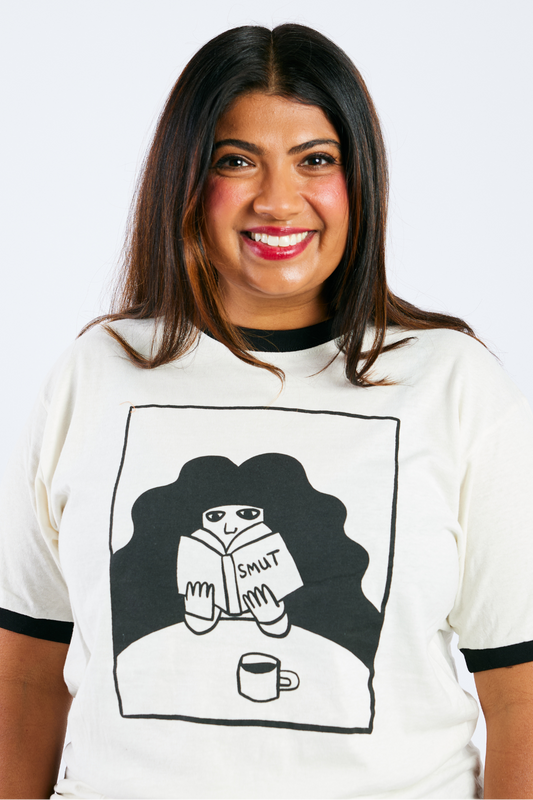 Woman wearing a white t-shirt with an illustrated woman reading smut and drinking coffee on a plain background