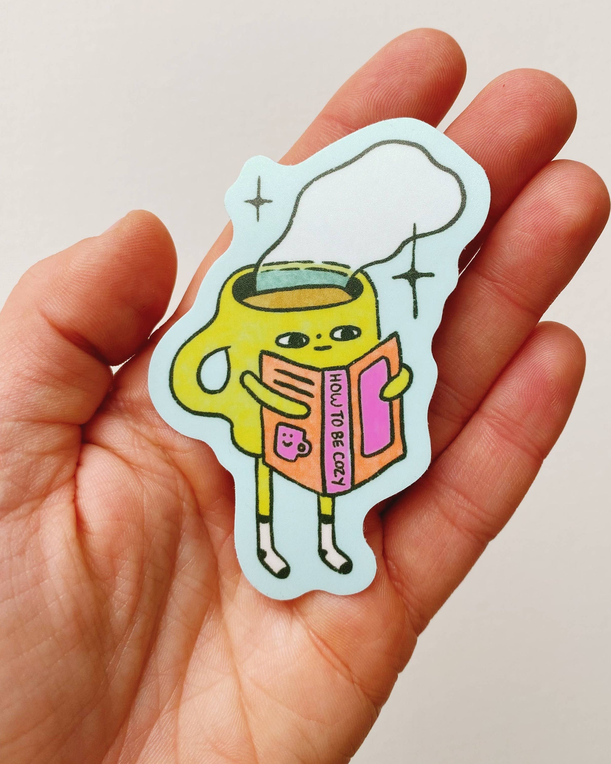 Hand holding a colorful sticker with a cartoon coffee mug holding a book titled how to be Cozy.  