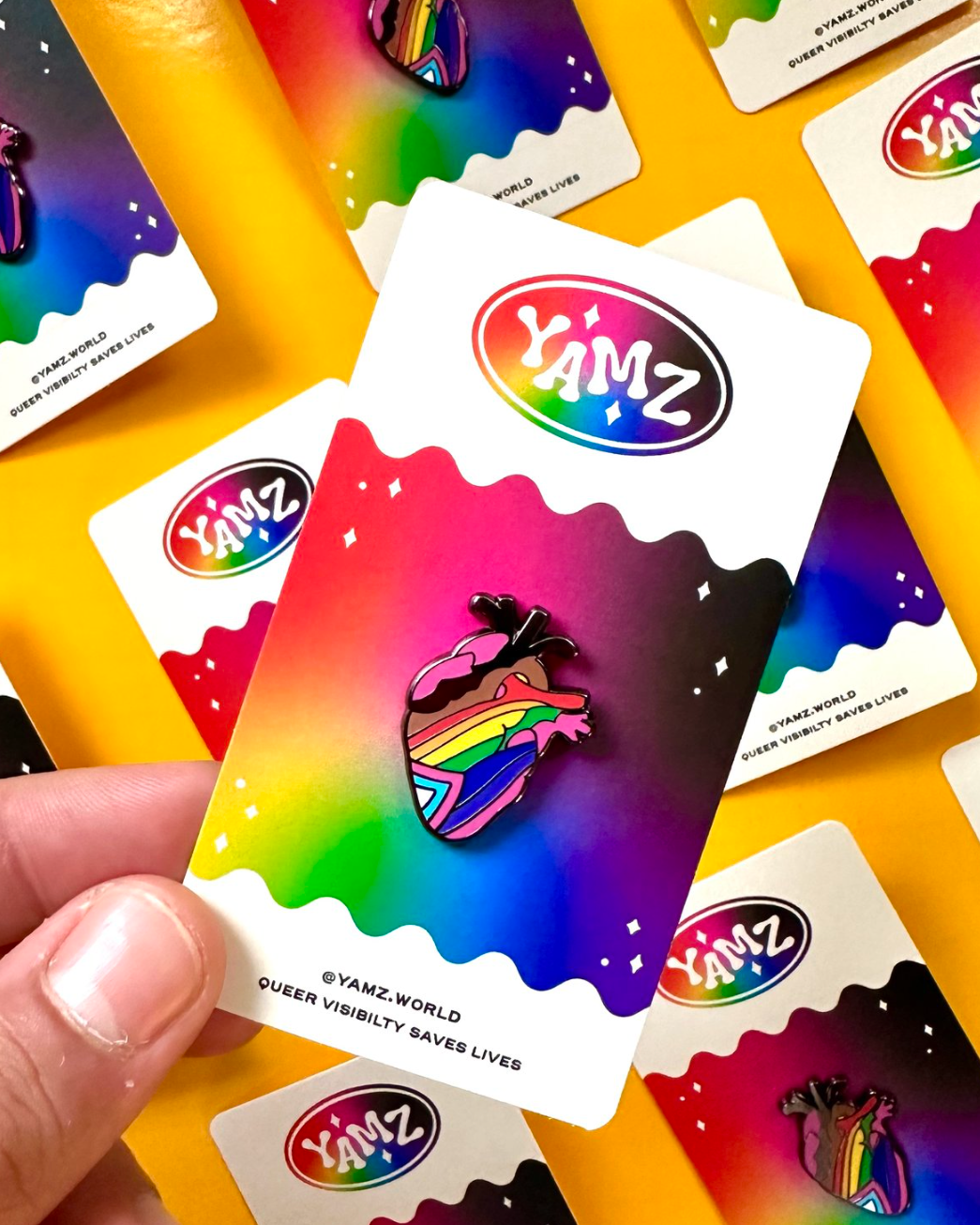 A hand holding a hard enamel pin with a colorful heart design on a card with the YAMZ logo.
