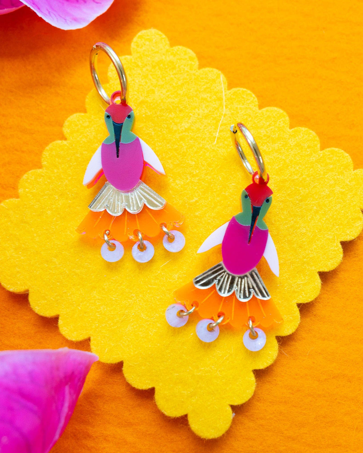 A pair of colorful hummingbird-shaped dangle earrings displayed on a yellow scalloped card over an orange background, with pink flower petals at the corners.