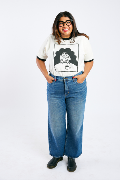 Person wearing a white ringer tee featuring an illustrated person reading a book labeled smut and blue jeans on a white background