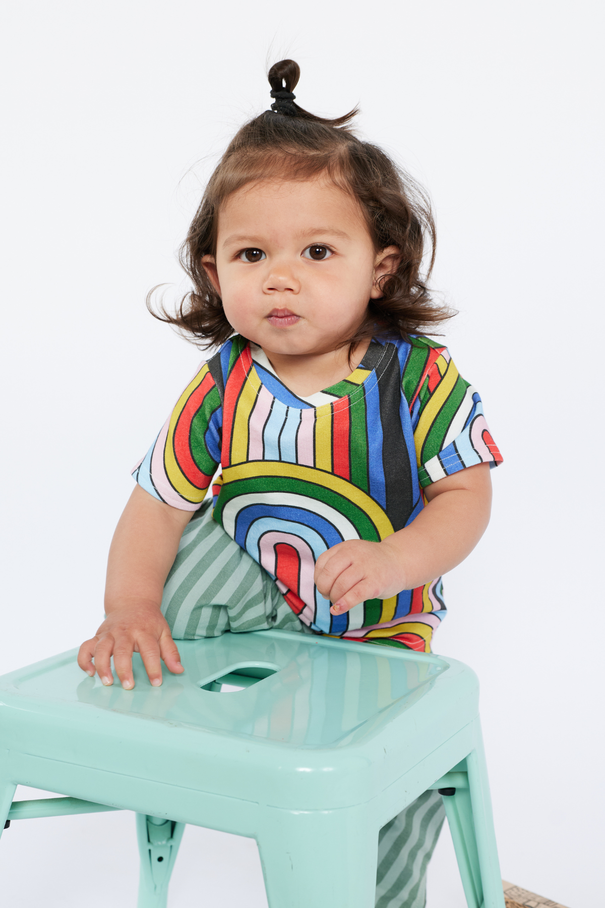 Child wearing a colorful organic cotton striped shirt sitting on a green stool against a white background
