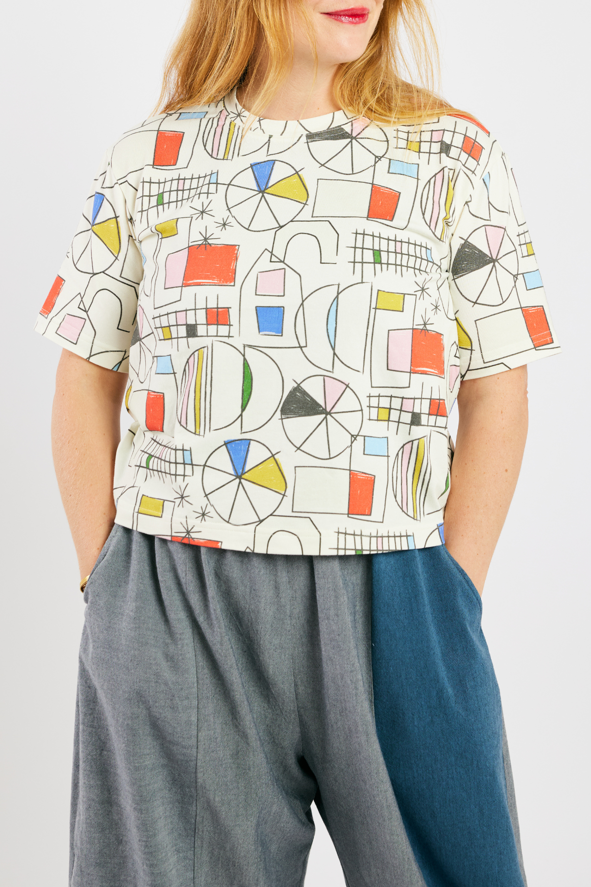 person with her hands in her pockets, wearing gray and blue barrel-leg pants made from sustainable bamboo and cotton. She's also wearing a colorful, organic cotton t-shirt.