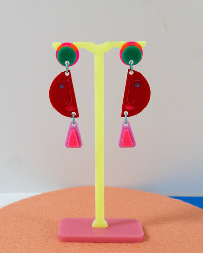 Colorful geometric drop earrings displayed on a yellow stand. The earrings feature red semi-circular dangles with pink triangular drops, accented by green and pink circular studs.