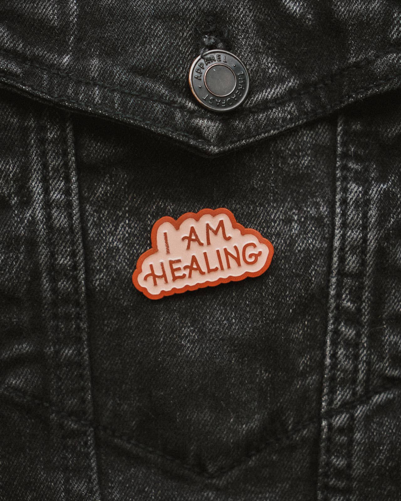 enamel pin with 'I AM HEALING' text on a black denim jacket
