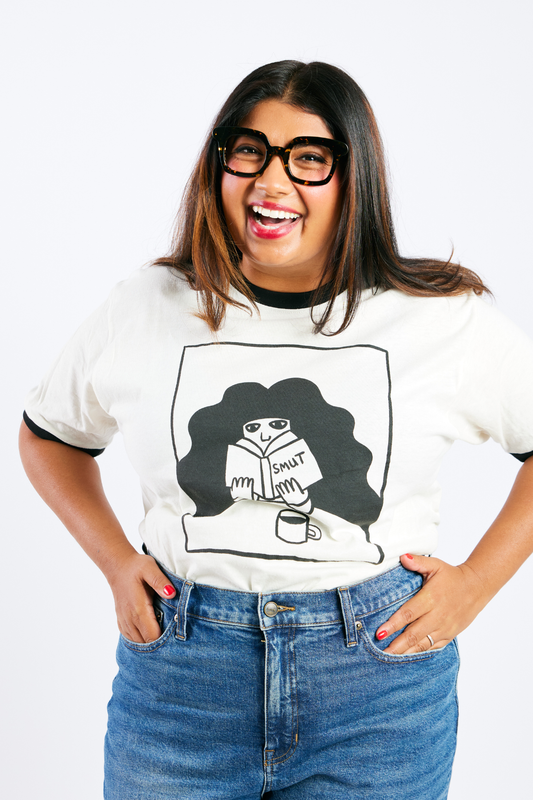 Person wearing a white t-shirt with an illustrated woman reading a smutty book and drinking coffee 