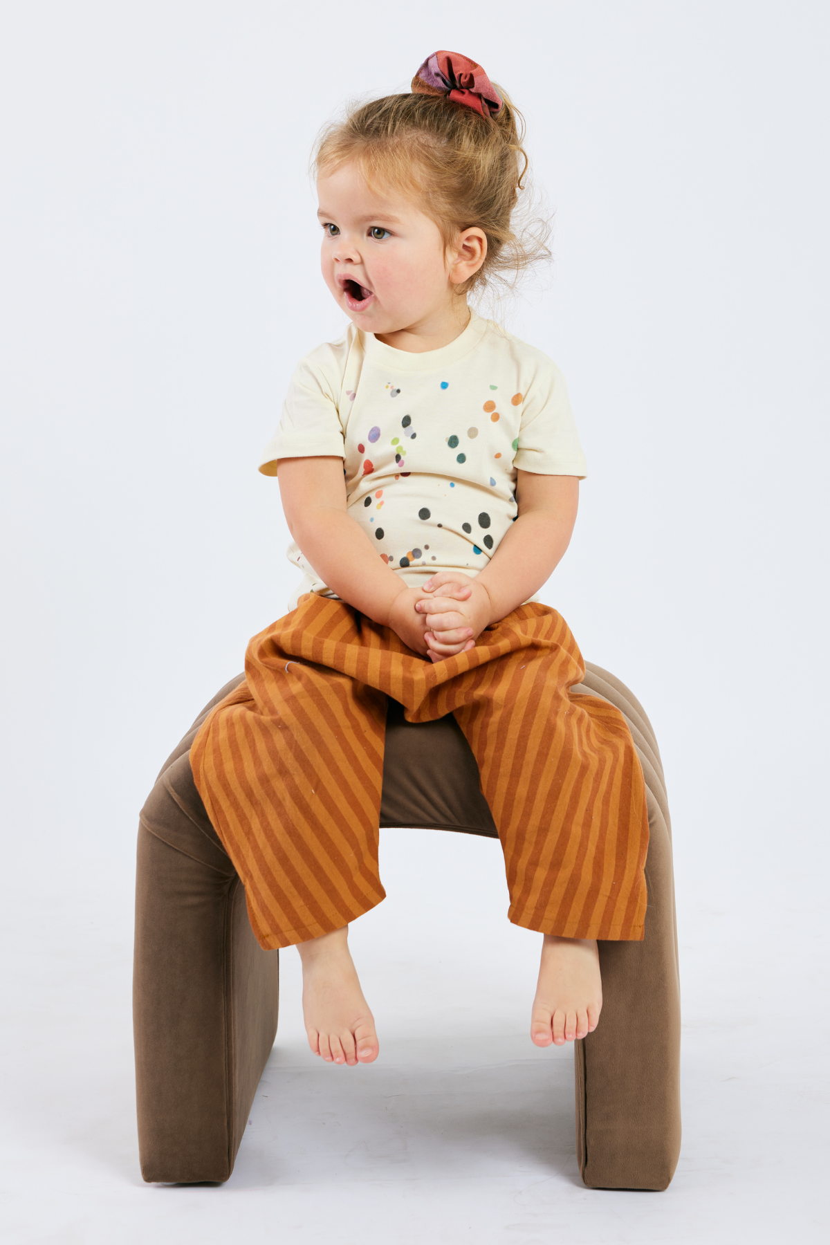 Child sitting on a brown cushioned stool and wearing a cream tshirt with colorful dots - against a white background