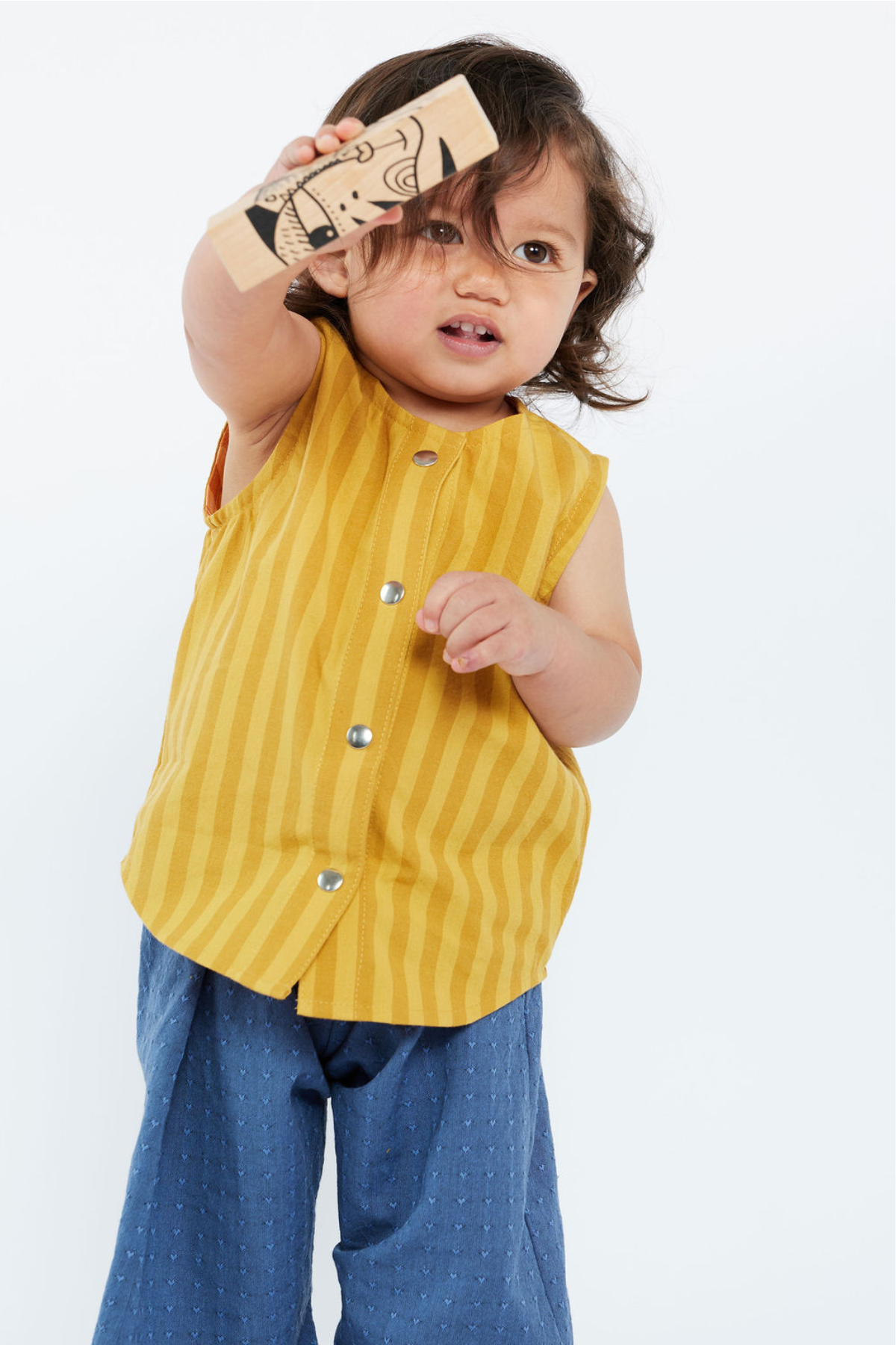 Child wearing a yellow sleeveless shirt with buttons and blue pants, holding a wooden toy.