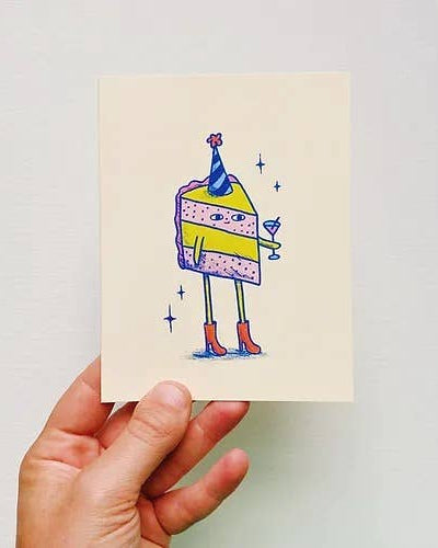 Hand holding a card with a colorful illustrated piece of cake in a party hat against a light gray background