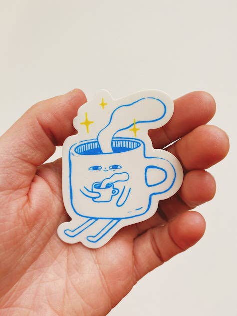 Hand holding a sticker of a cartoon cup with a face and steam rising from it.