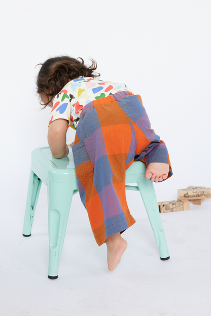 Child climbing on a green stool wearing colorful pants and a patterned shirt on a white background