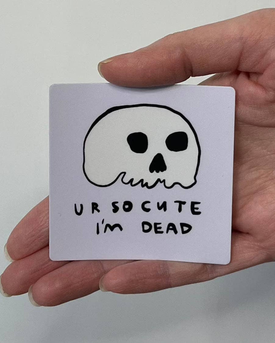 Hand holding a sticker with a skull drawing and text 'UR SO CUTE I'M DEAD' on a plain background